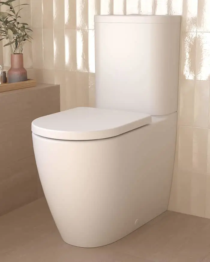 White modern toilet in a bathroom with beige tiles, sheer curtains, and a plant in a vase on a shelf.