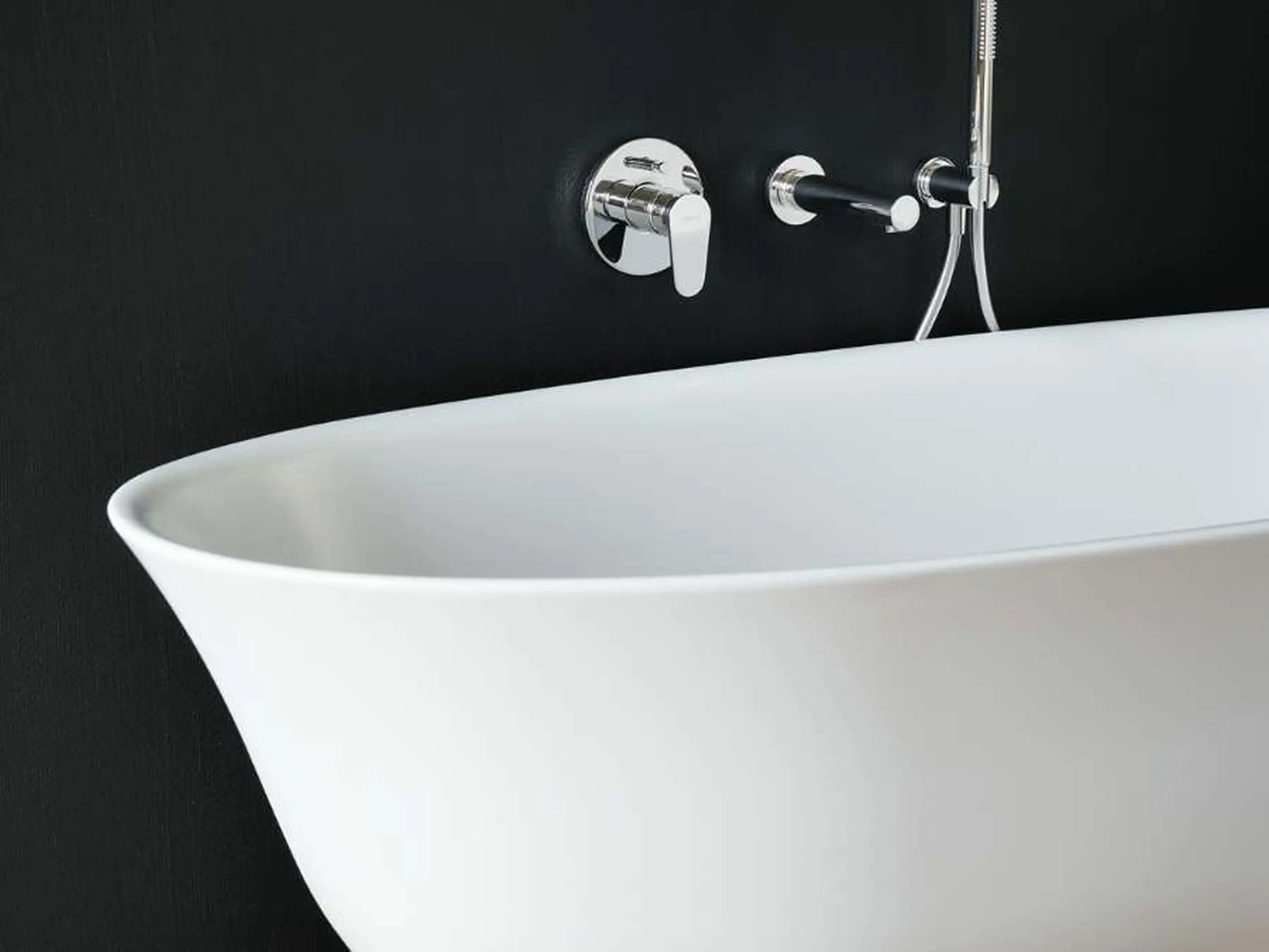 Modern white freestanding bathtub against black wall with chrome shower fixtures and handheld sprayer.