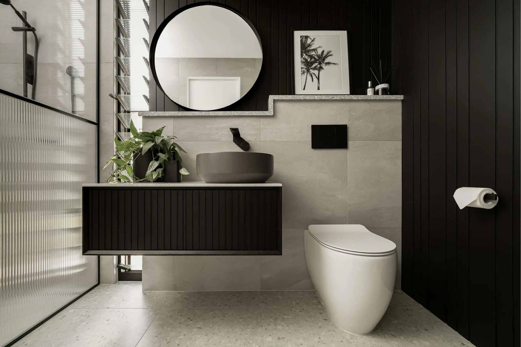 Modern bathroom with black and gray design featuring round mirror, vessel sink, white toilet, and potted plant.