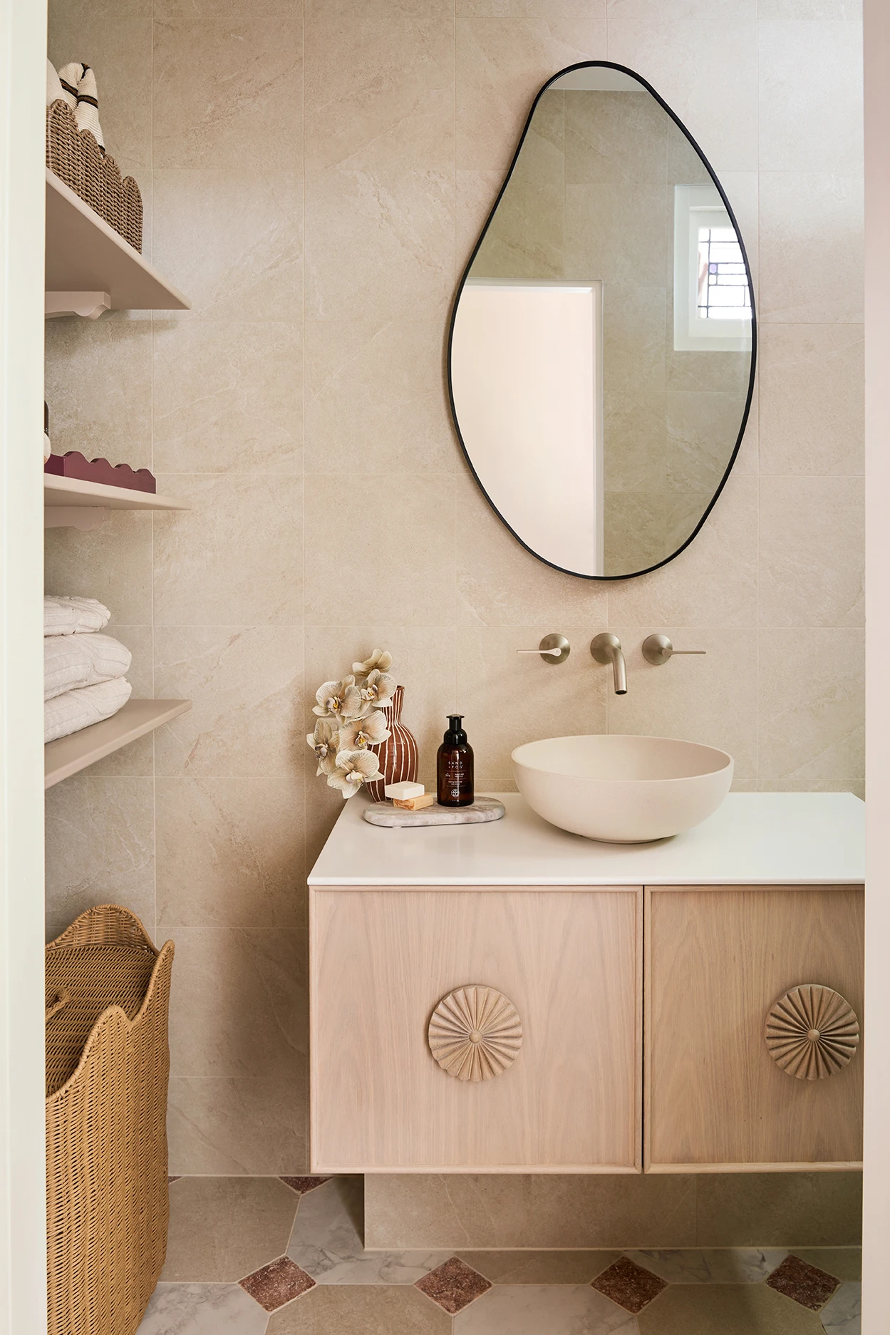Bathroom with a floating wood vanity, bowl sink, oval mirror, beige tiles, open shelves with towels, and a wicker basket.