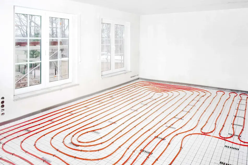 Empty white room with large windows showing installed underfloor heating pipes in red and orange arranged in loops.