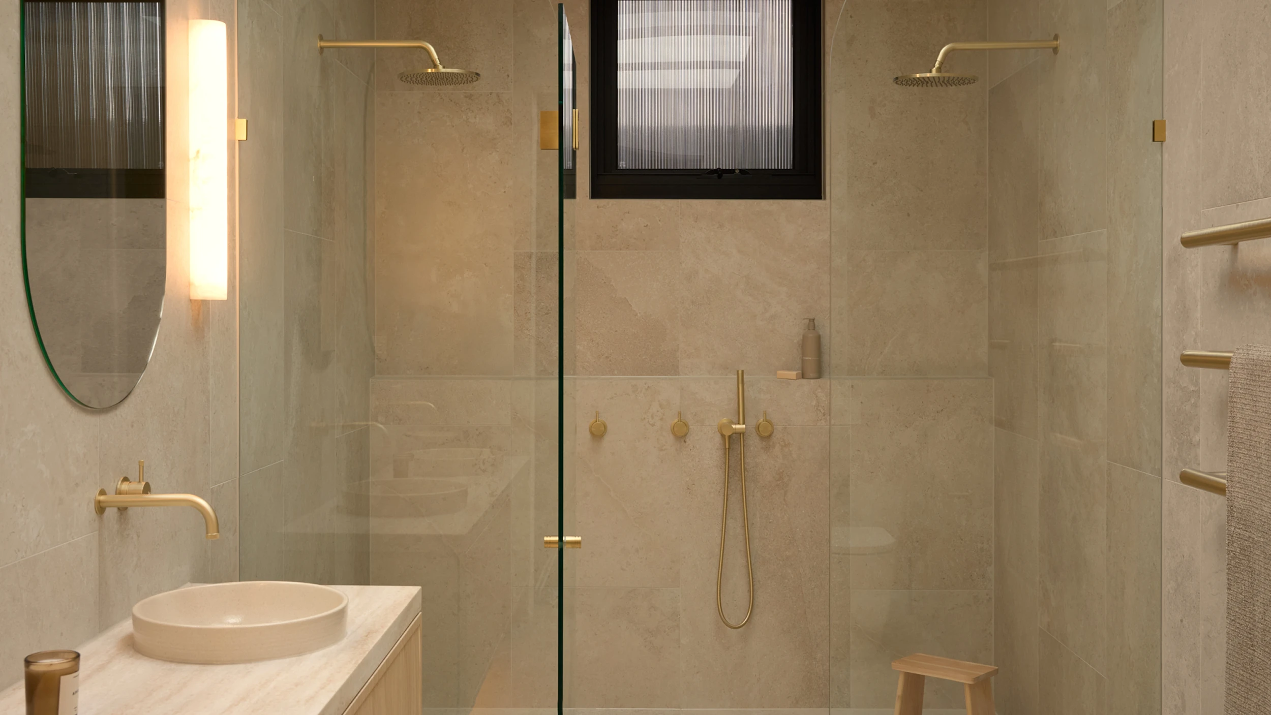 Modern bathroom with beige tile walls, glass shower enclosure, gold fixtures, round sink, and vertical light fixture.