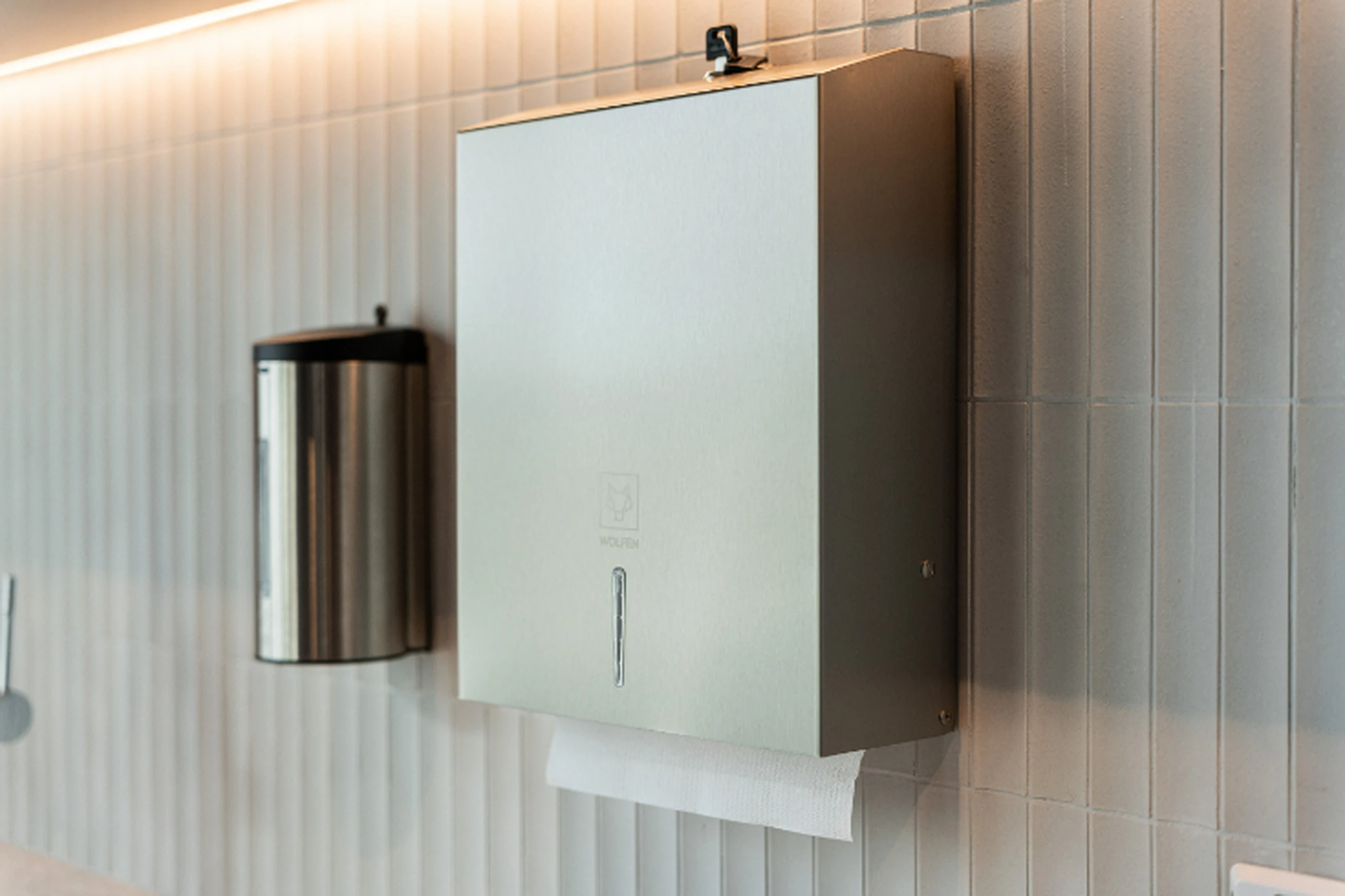 Modern paper towel dispenser and stainless steel waste bin mounted on light-colored tiled bathroom wall.