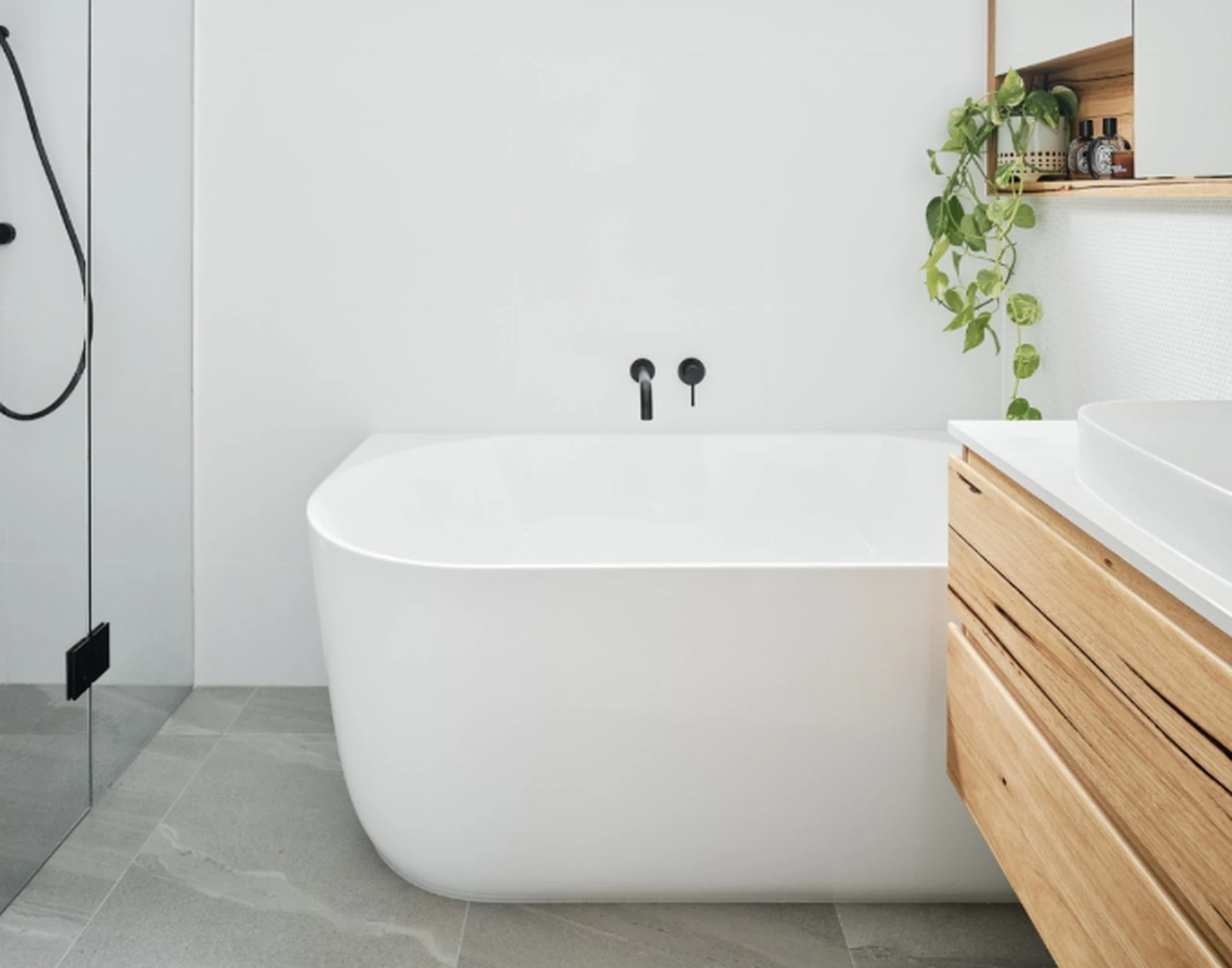 Modern white bathroom with freestanding tub, wooden vanity, black fixtures, and trailing plants on a shelf.