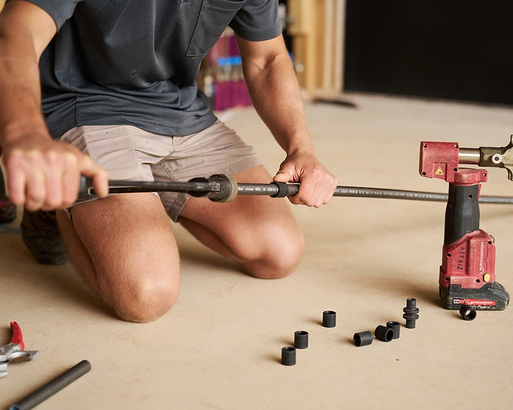 Person in gray shirt and beige shorts kneeling on floor, using tools with a red power drill nearby.