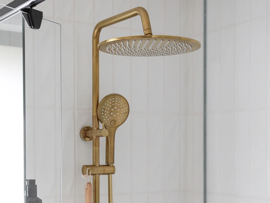 Modern brass shower system with large rainfall showerhead and handheld sprayer mounted on white tiled bathroom wall.