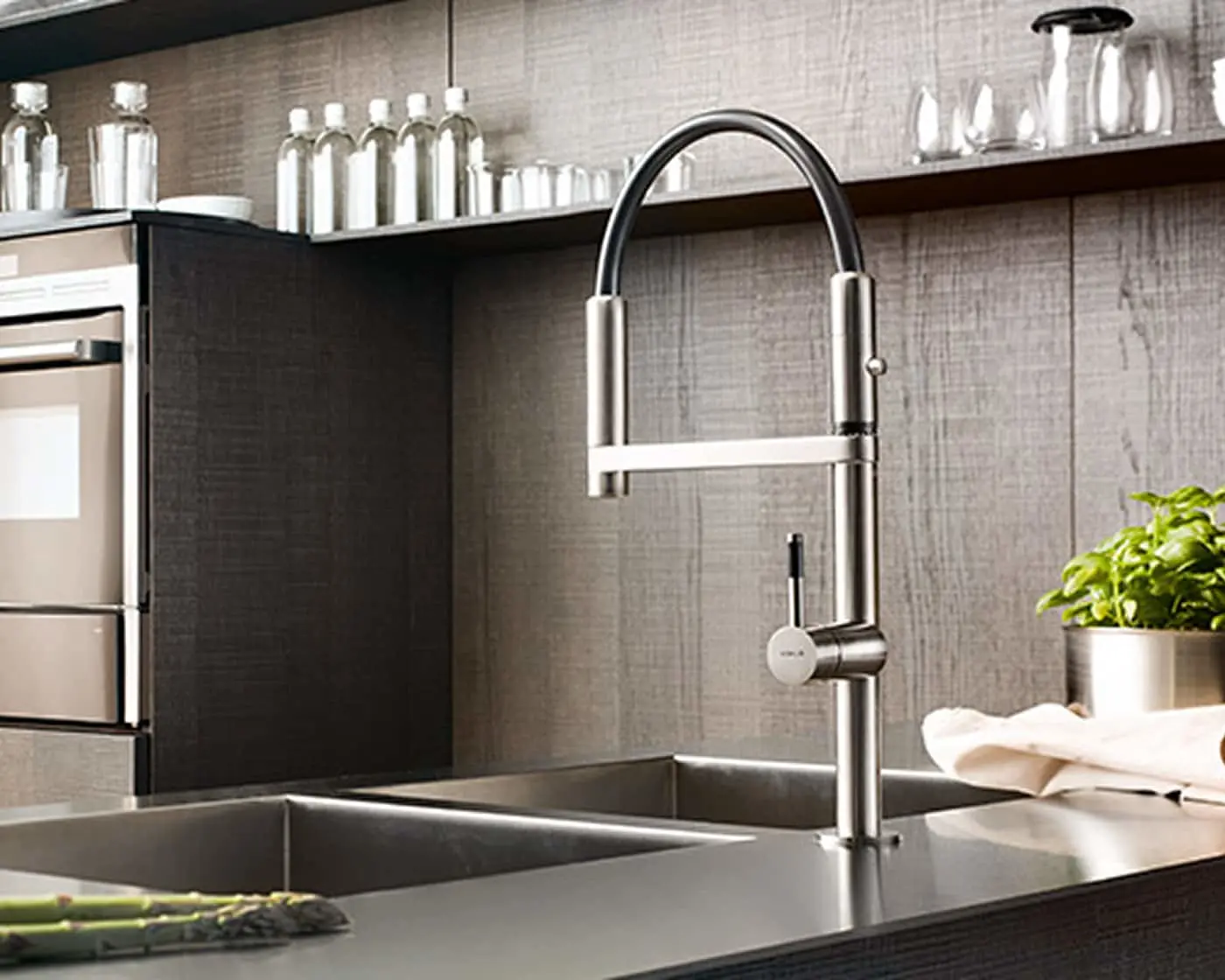 Modern stainless steel kitchen faucet with pull-down sprayer over a double sink, with glass bottles on shelves above.