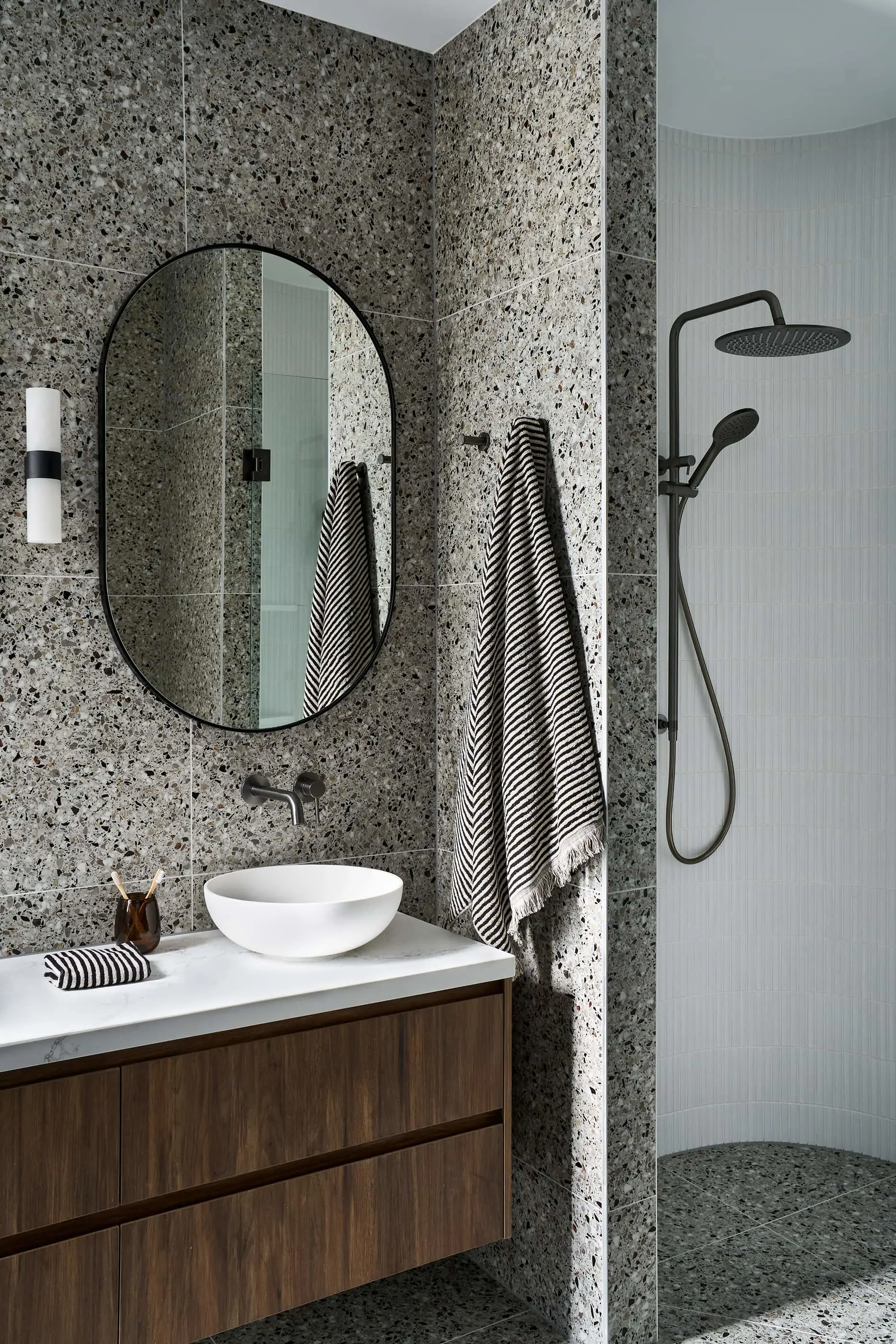 Modern bathroom with terrazzo walls, oval mirror, wooden vanity, white basin, striped towels, and walk-in shower.