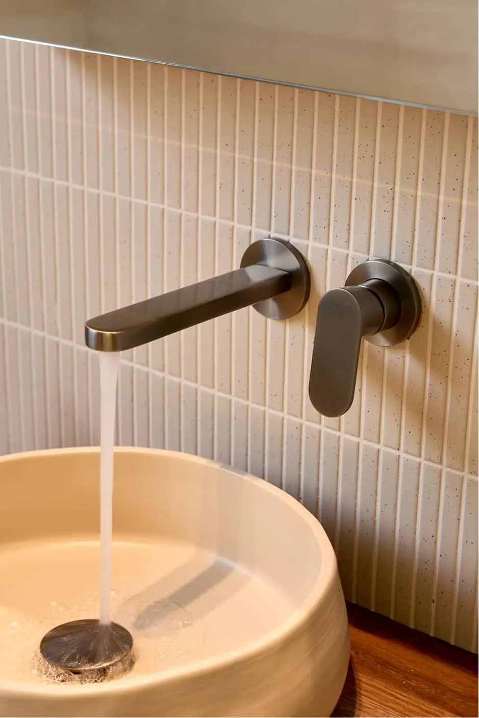 Wall-mounted gunmetal faucet with water flowing into a round white sink against beige vertical tile backsplash.