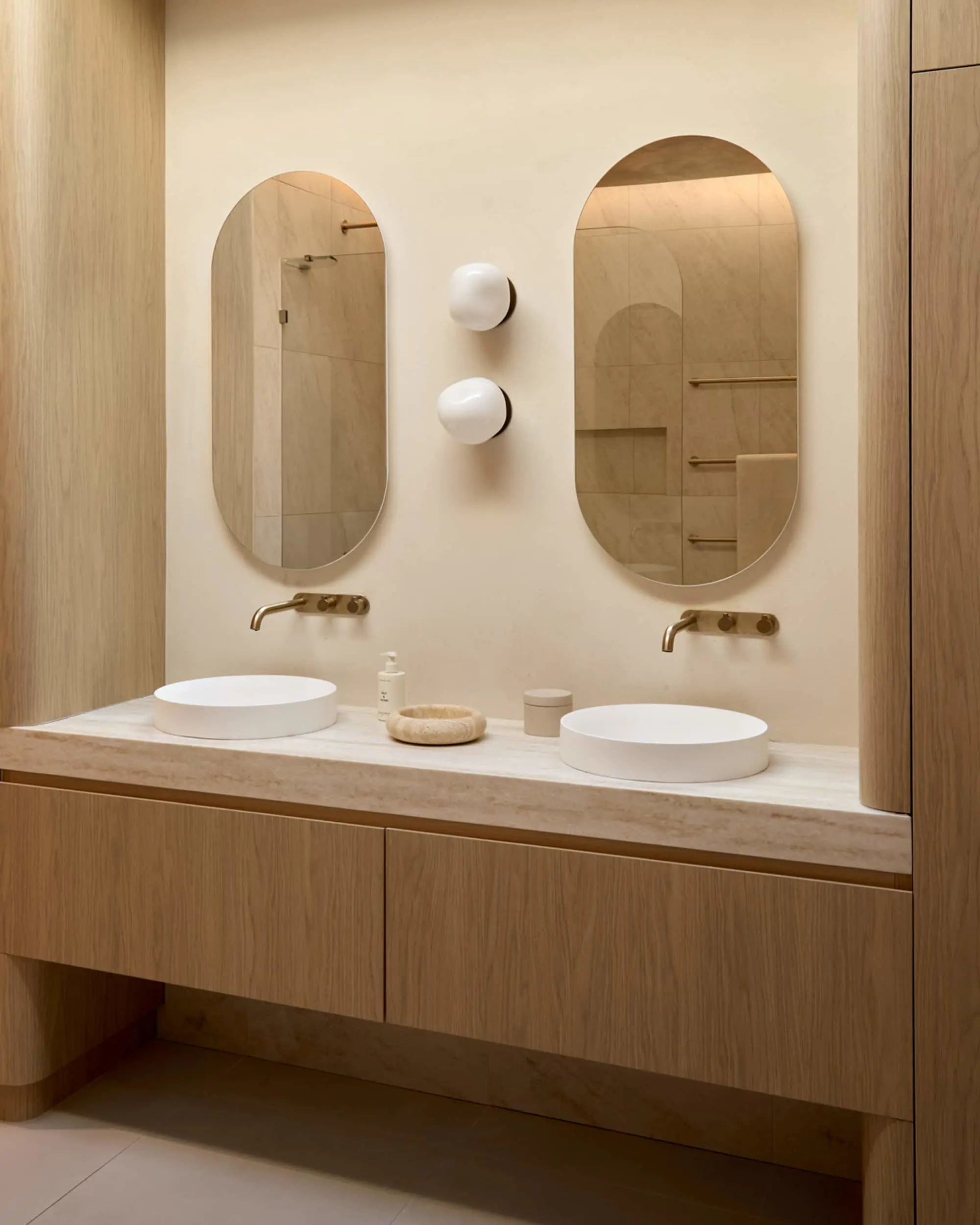 Bathroom vanity with two white vessel sinks on a stone countertop, wood cabinetry, oval mirrors, and two white globe wall lights.