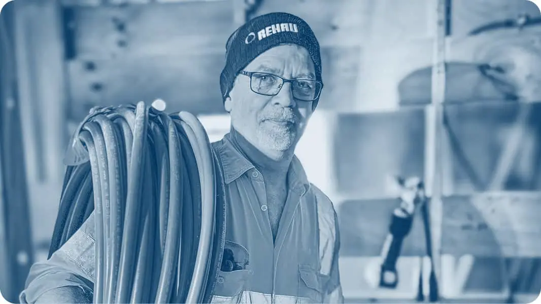 Worker in blue-tinted image carrying coiled pipes or tubing over shoulder, wearing REHAU branded knit cap at construction site.