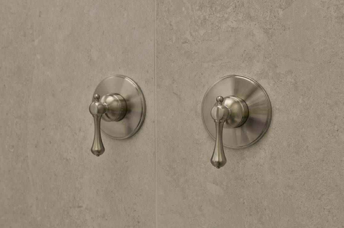 Two brushed nickel shower handles with teardrop-shaped levers mounted on beige stone tile wall.