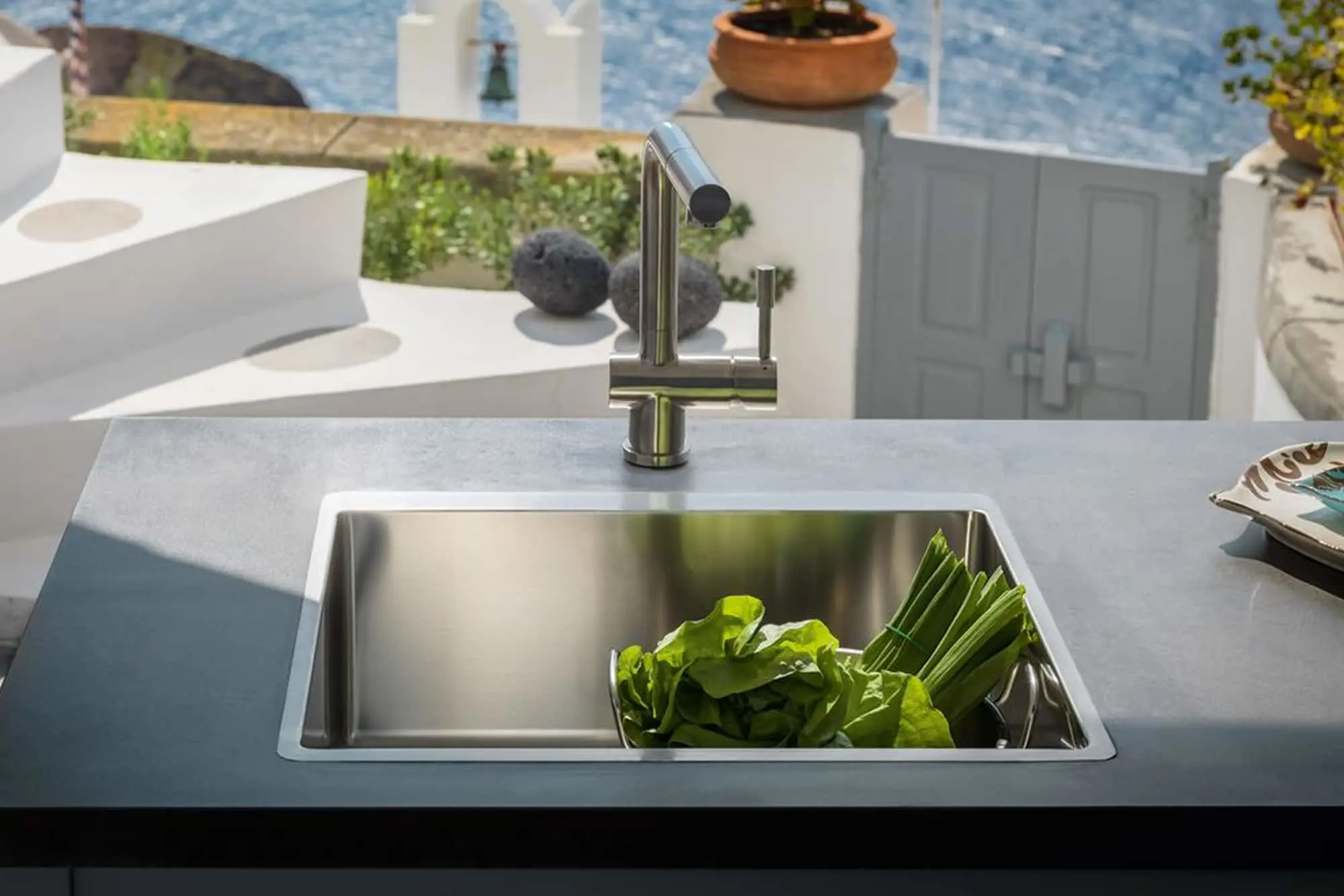 Modern outdoor kitchen sink with fresh greens, overlooking Mediterranean sea with white architecture and blue water.