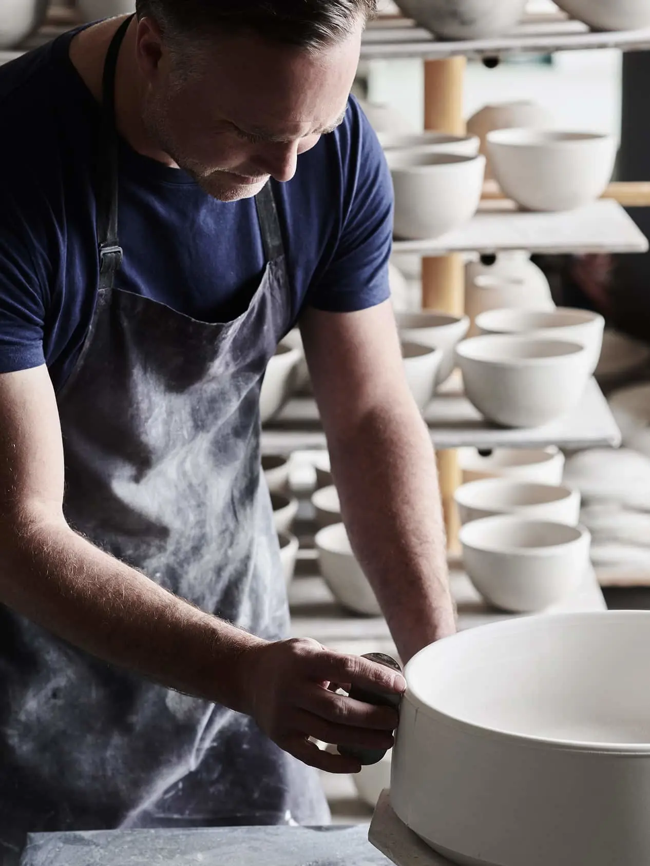 Potter in blue shirt and gray apron crafting a white ceramic bowl with shelves of pottery in background.