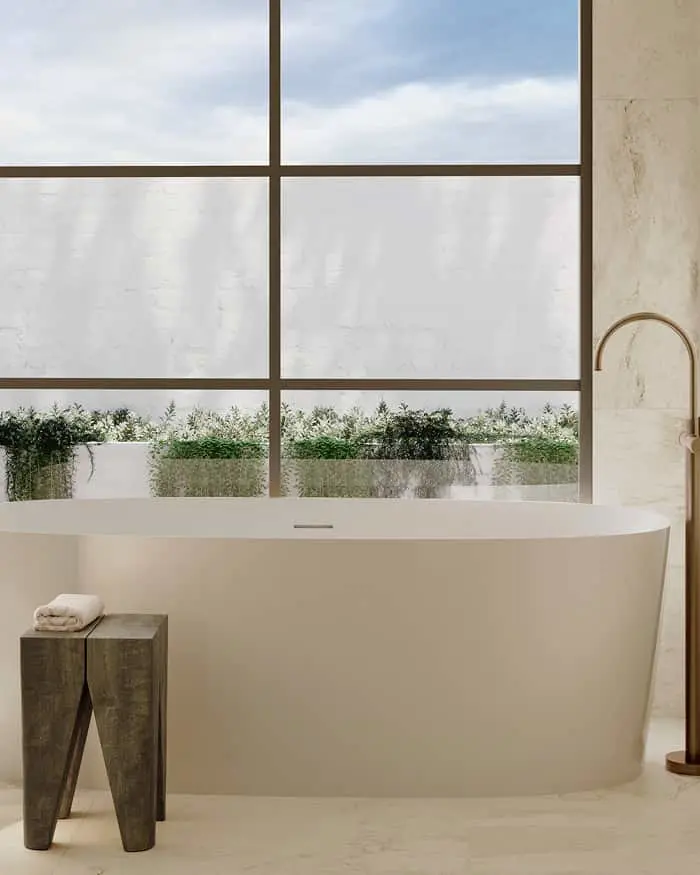 Modern white freestanding bathtub next to wooden stool in marble bathroom with large windows overlooking greenery.