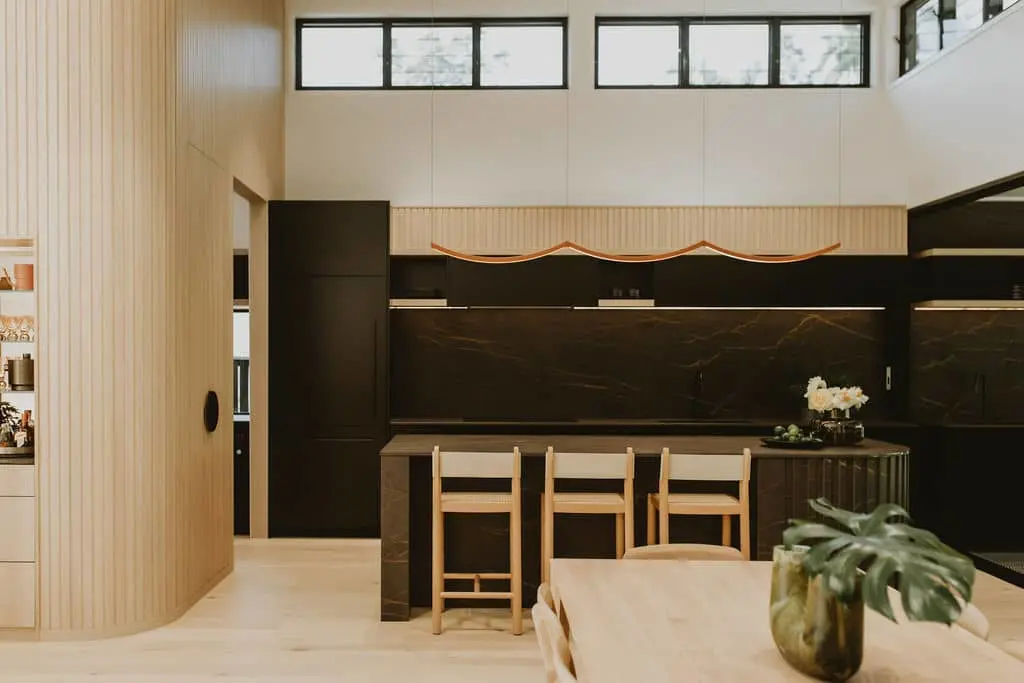 Modern kitchen with black marble backsplash, wooden bar stools, light wood paneling, and high windows.