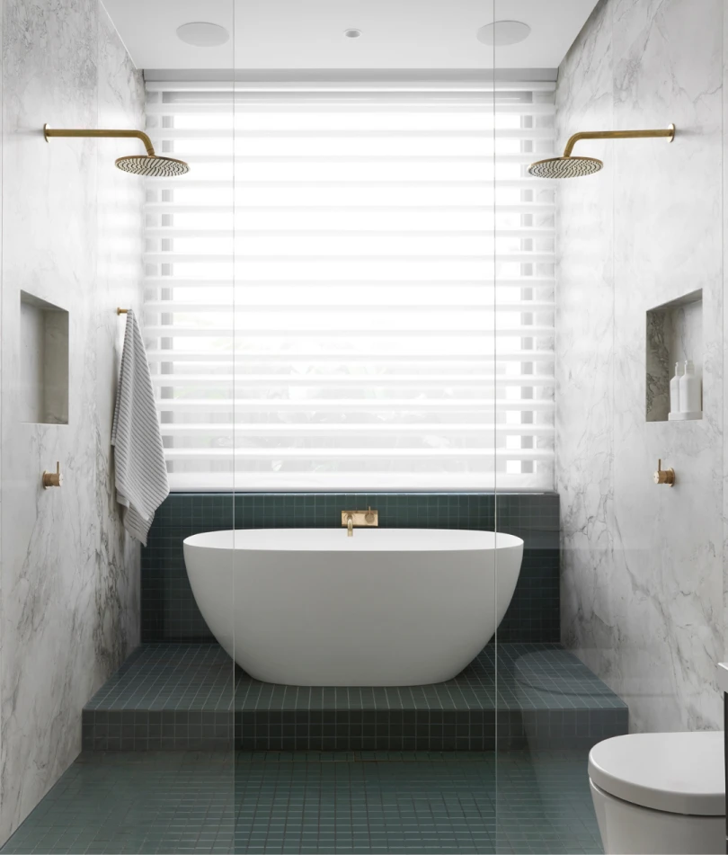Modern bathroom with white freestanding tub on green tile platform, marble walls, brass fixtures, and window blinds.