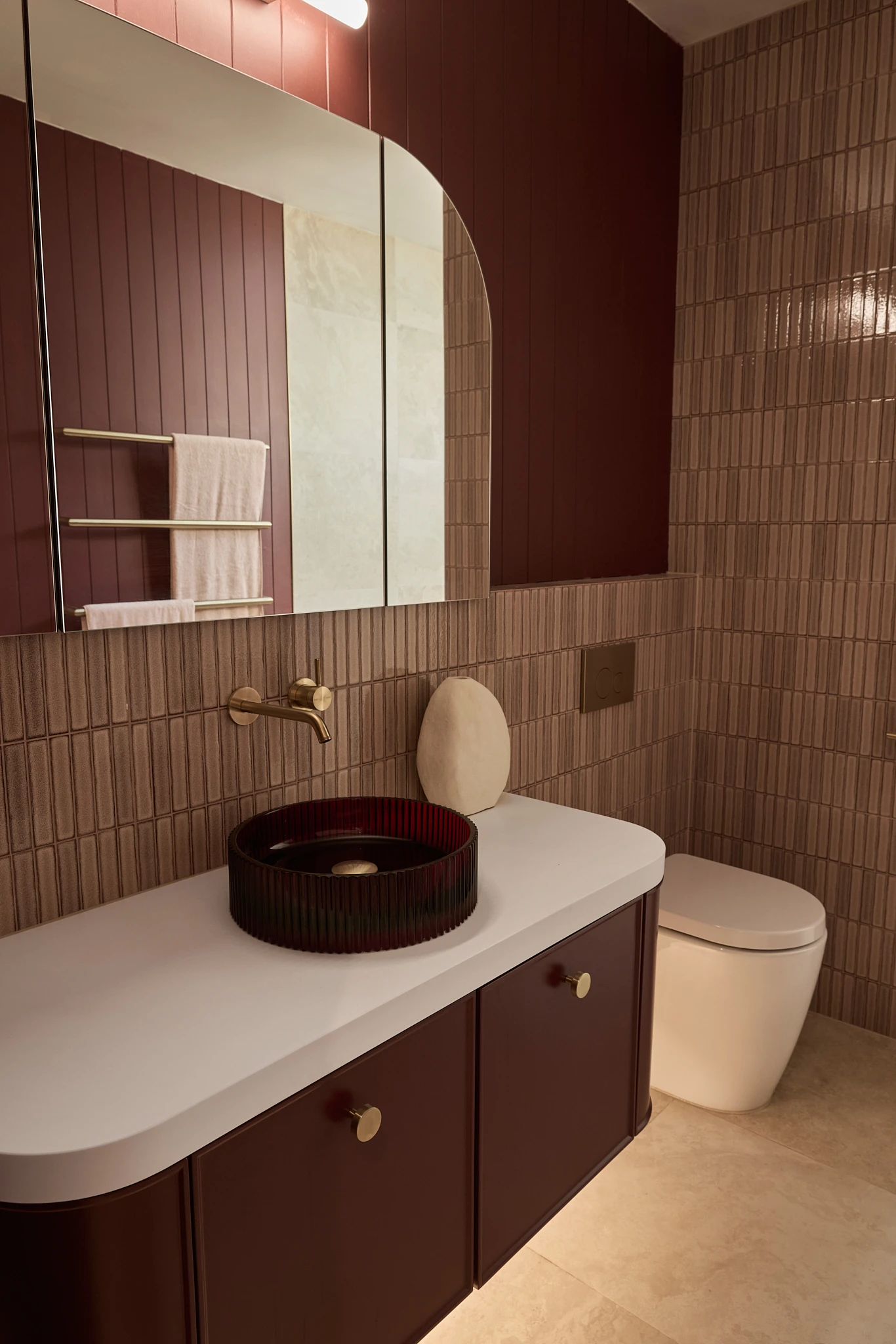Modern bathroom with burgundy cabinetry, beige tile walls, white countertop, dark red vessel sink, and gold fixtures.