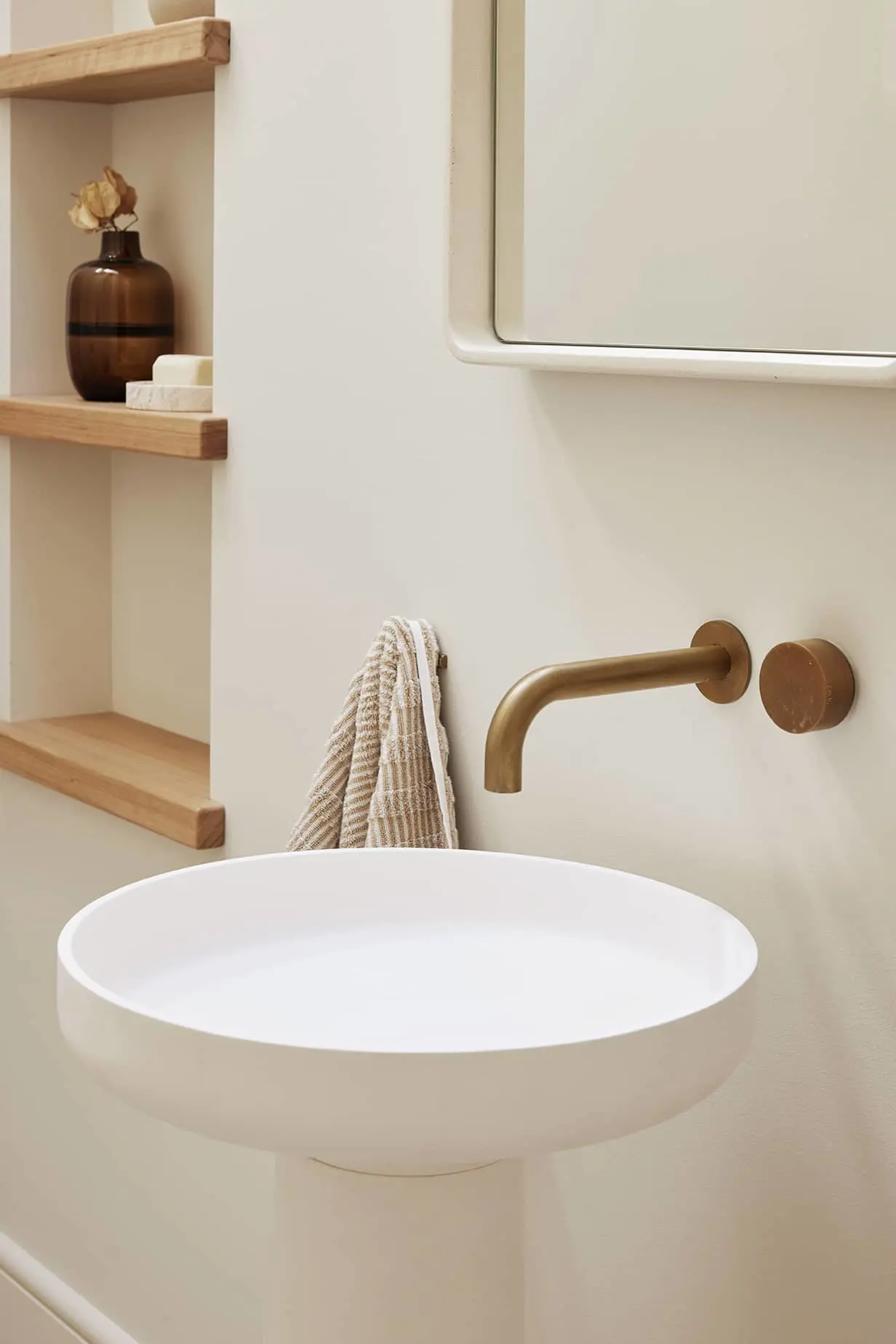 Modern bathroom with white basin sink, brass wall-mounted faucet, wooden shelves with brown vase, and striped towel.