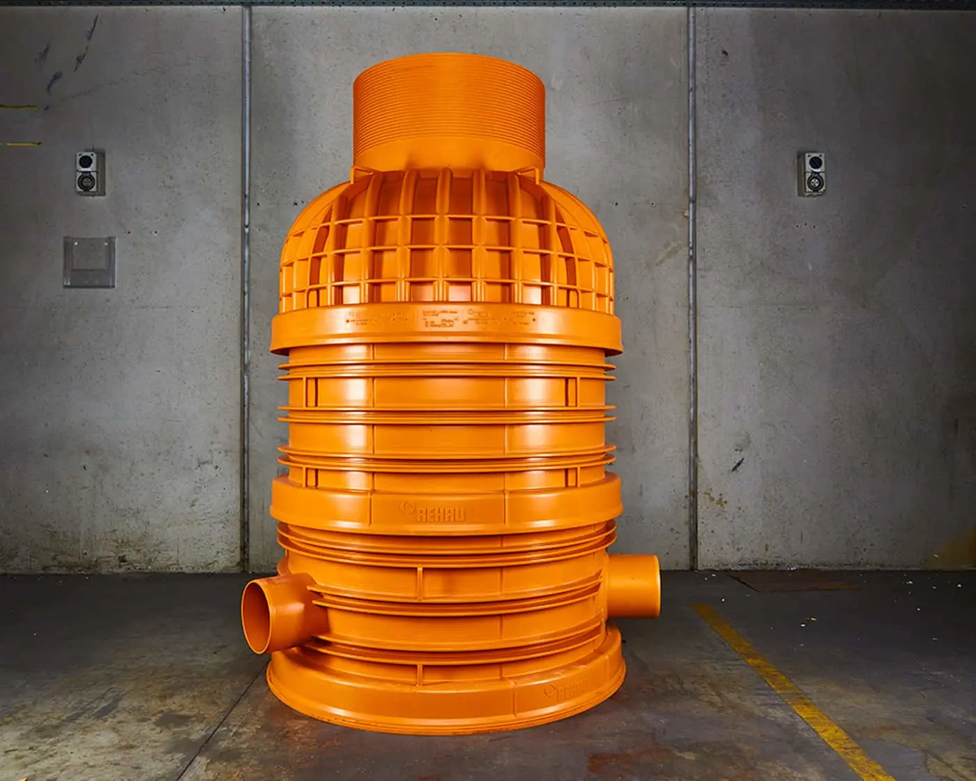 Large orange industrial pipe fitting with ribbed design against concrete wall in warehouse setting.