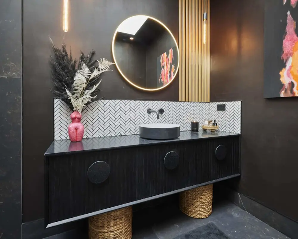 Modern bathroom with dark walls, round backlit mirror, herringbone tile backsplash, and pink vase with dried plants.
