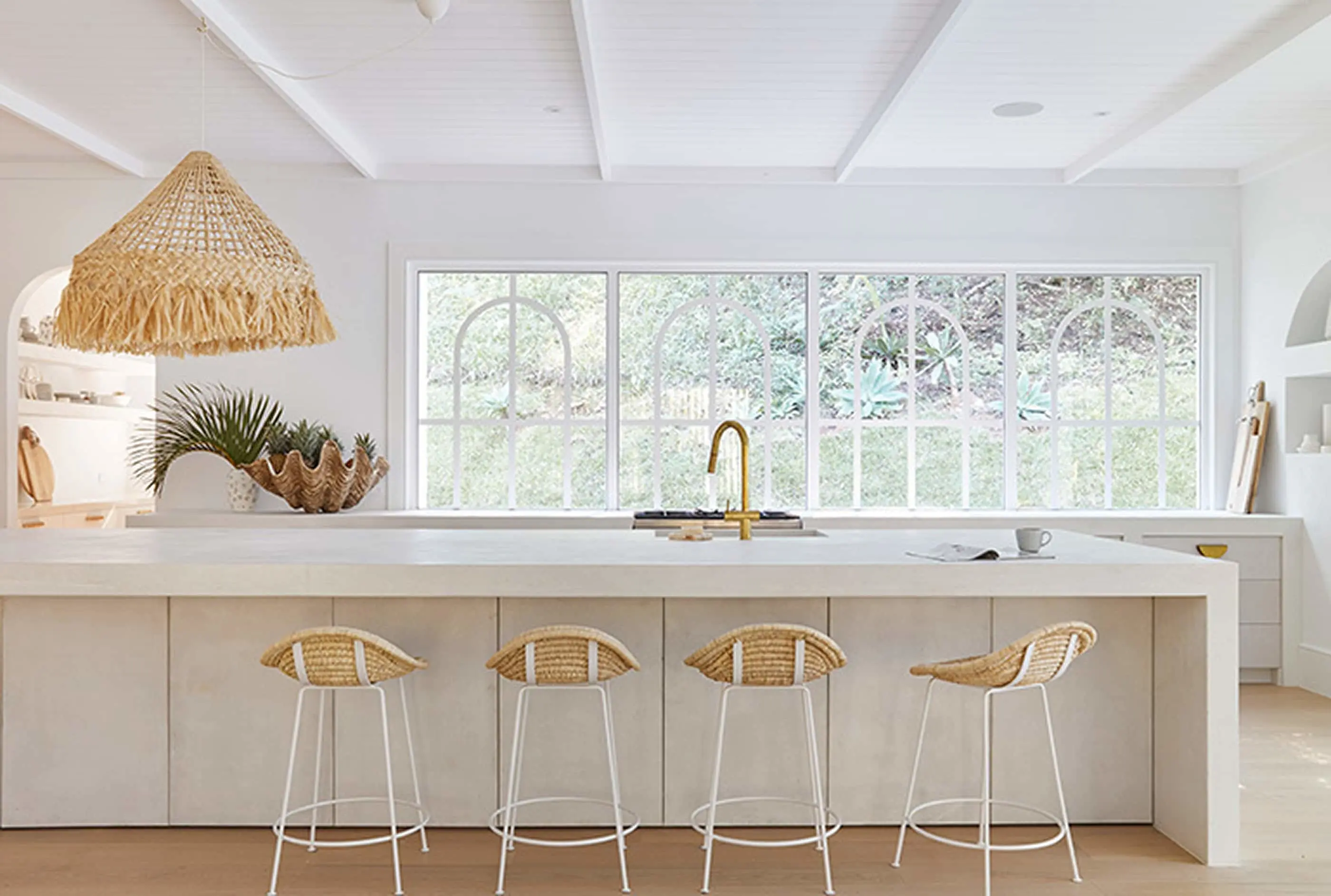 Bright white kitchen with woven pendant light, gold faucet, four rattan barstools, and large windows overlooking greenery.