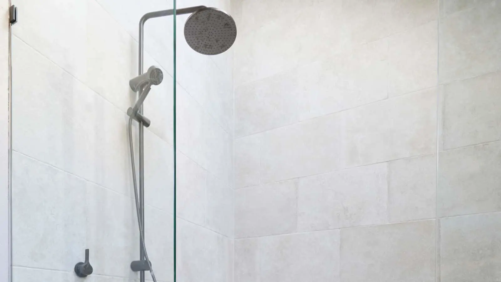 Modern shower with rainfall showerhead, handheld sprayer, and glass partition in a white tiled bathroom.