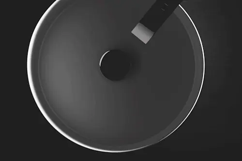 Overhead view of a minimalist round sink basin with a black faucet against dark background in monochrome.