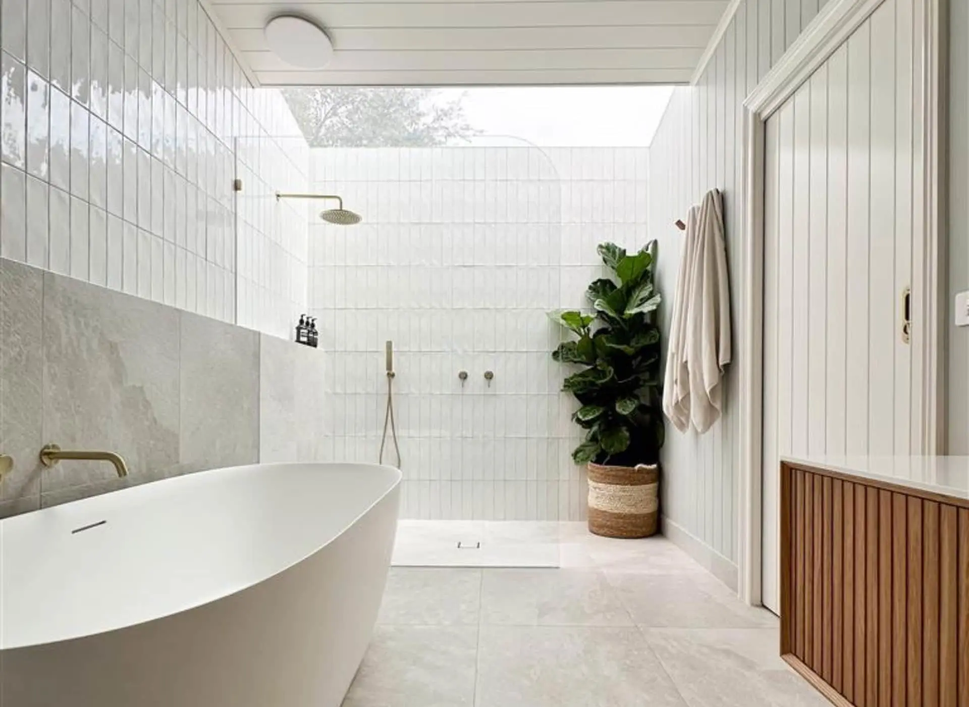 Modern white bathroom with freestanding tub, walk-in shower, skylight, and potted plant in woven basket.