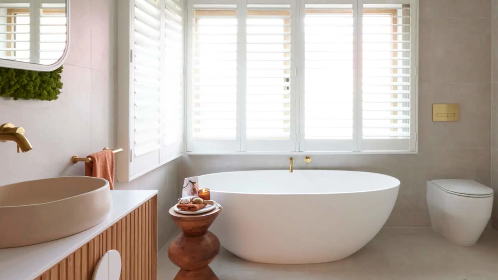 Modern bathroom with white freestanding tub, wooden vanity, beige sink, and large windows with white shutters.