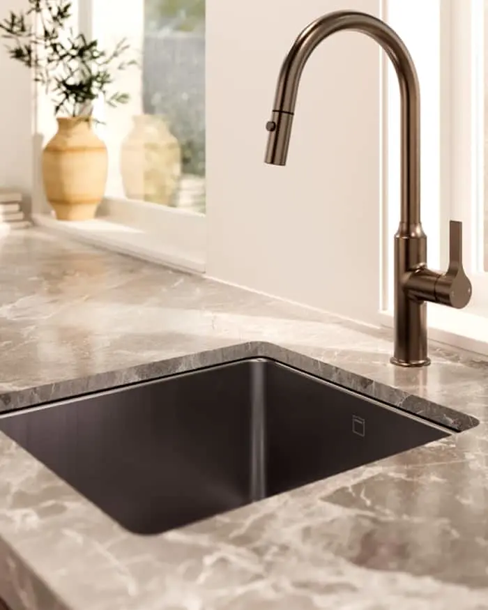 Modern kitchen sink with bronze pull-down faucet and dark basin set in marble countertop, ceramic vase in background.