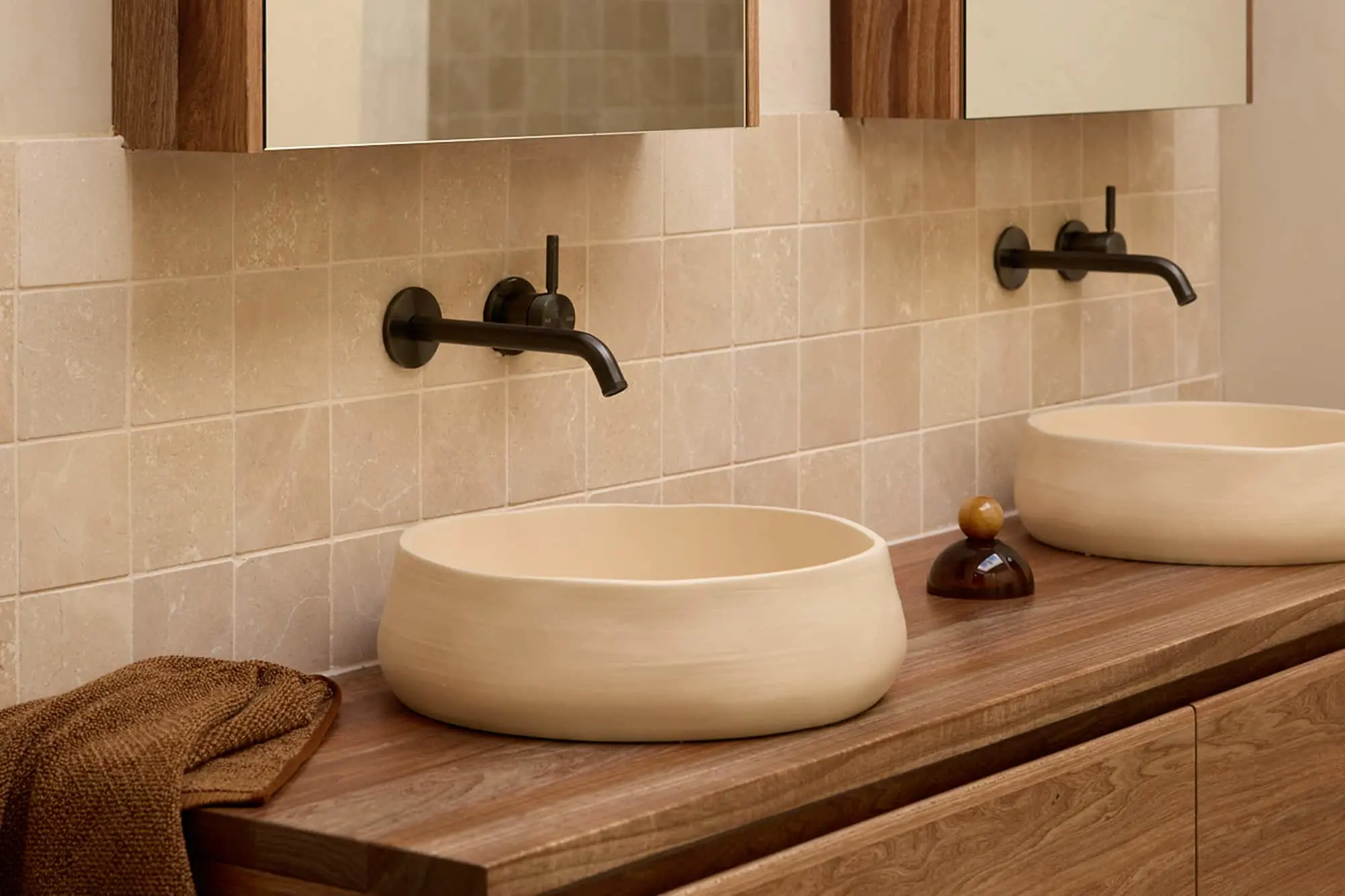 Two cream-colored vessel sinks on a wooden vanity with black wall-mounted faucets and beige tile backsplash.