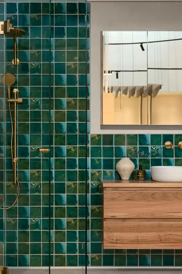 Bathroom with teal square tiles, brass shower fixtures, a wood vanity with white basin, decorative vase, and a wall mirror.