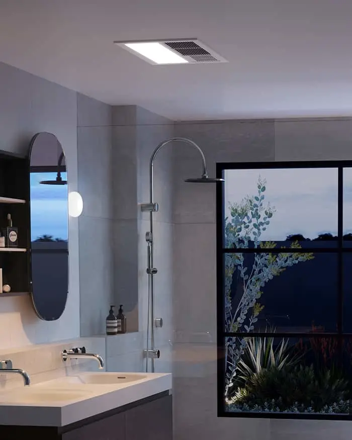 Modern bathroom with rainfall shower, oval mirror, white sink, and large window showing plants and evening sky.