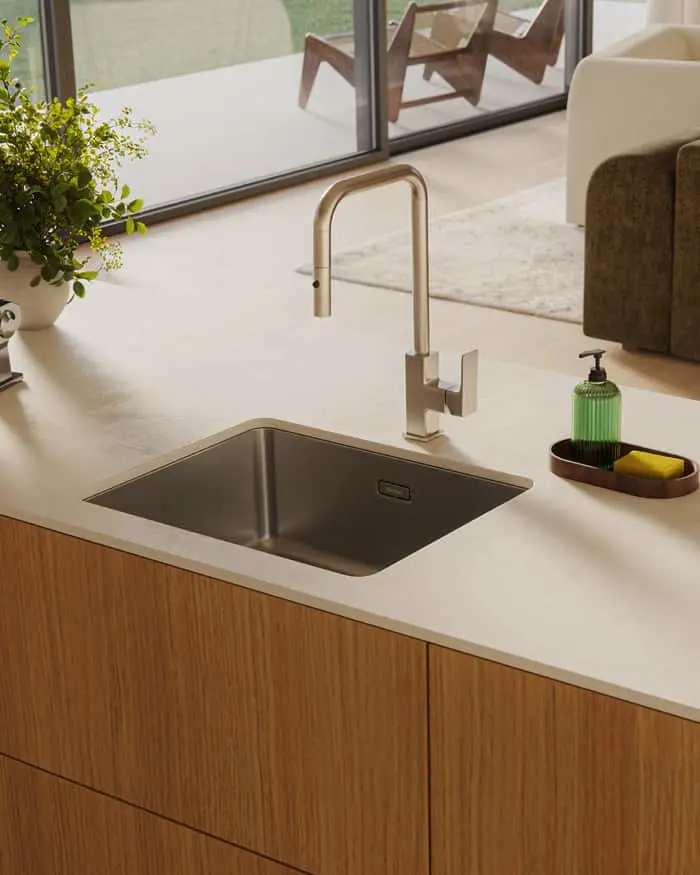 Modern kitchen sink with gold faucet on white countertop, wooden cabinets, and view of outdoor seating through window.