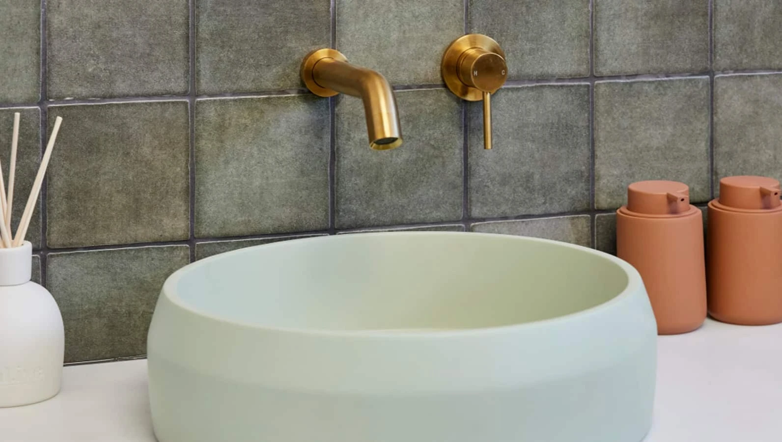Modern bathroom with white vessel sink, brass wall-mounted faucet, green tile backsplash, and terracotta dispensers.