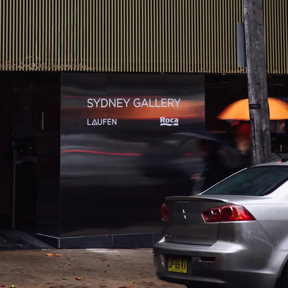 Sydney Gallery storefront with LAUFEN and Roca logos, silver car parked in front, and blurred person walking past.