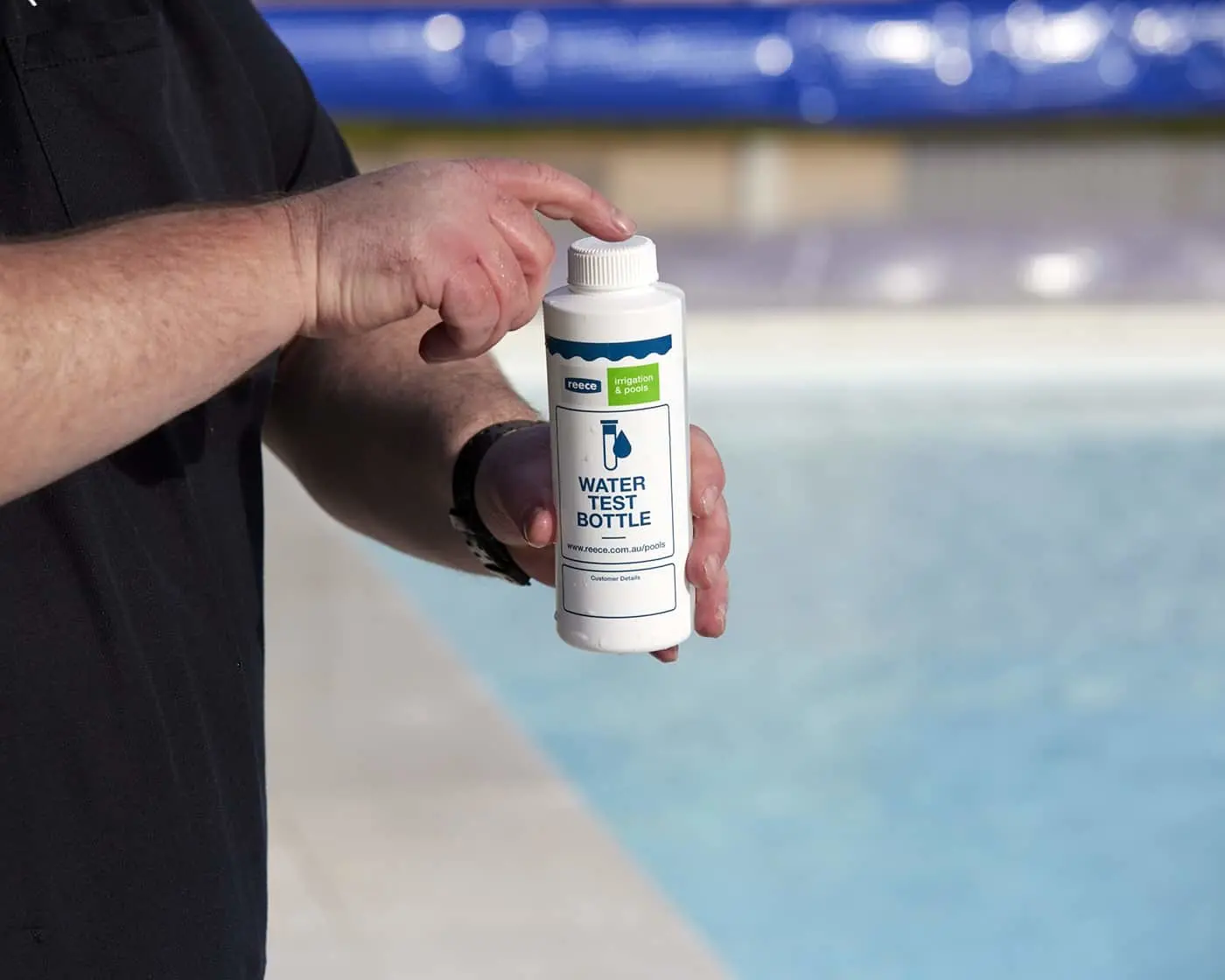 Hand holding a white Reece water test bottle beside a swimming pool with blue water in the background.