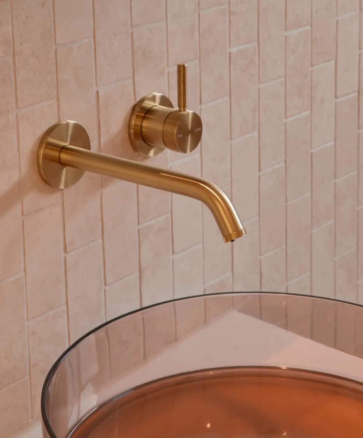 Brass wall-mounted faucet with separate control knob above a round sink against pink tiled bathroom wall.