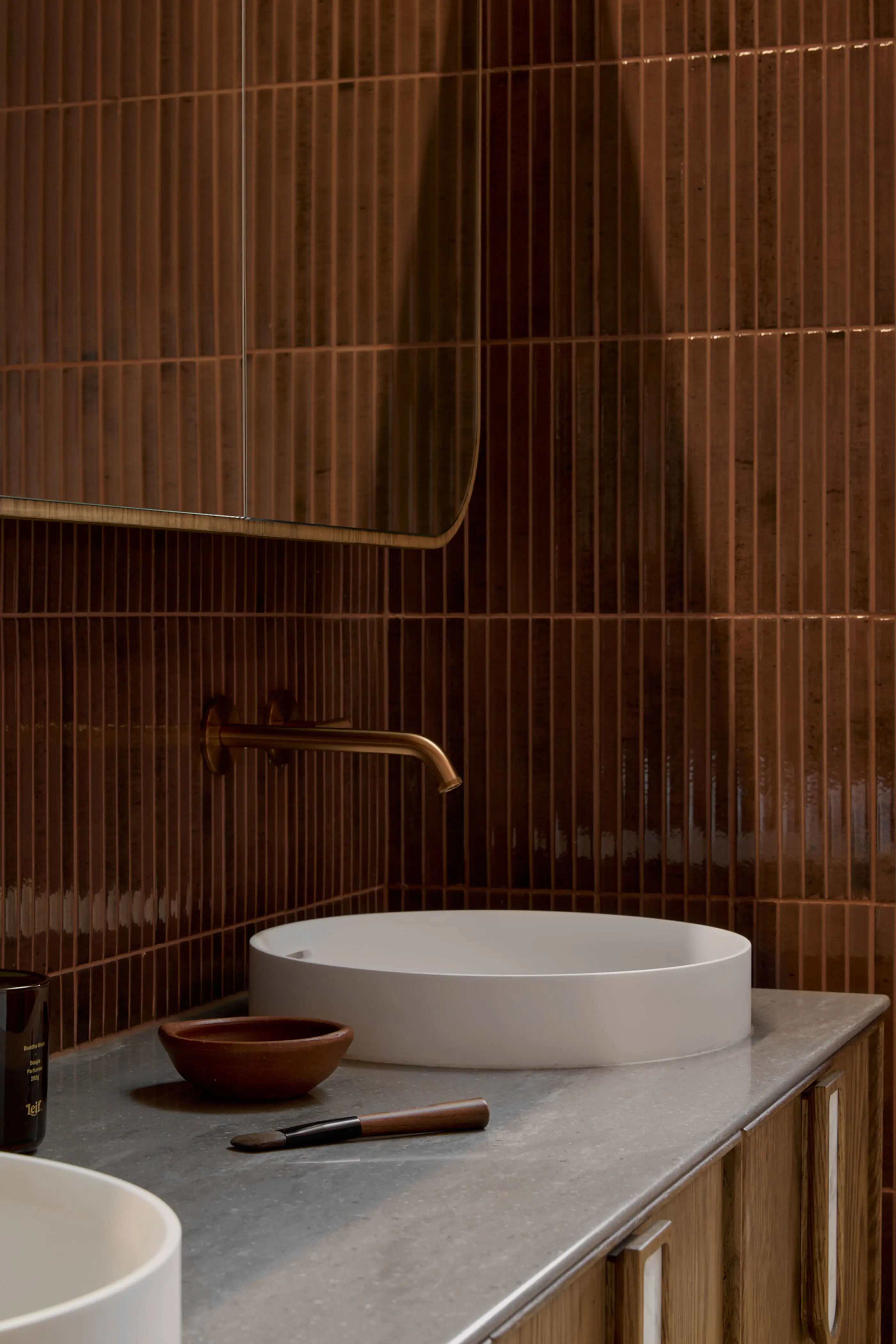 Modern bathroom with brown tiled walls, white circular sink, brass faucet, and wooden accents on a gray countertop.
