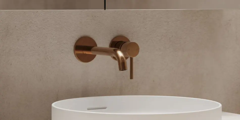 Modern copper wall-mounted faucet above a white basin sink against a beige textured wall.