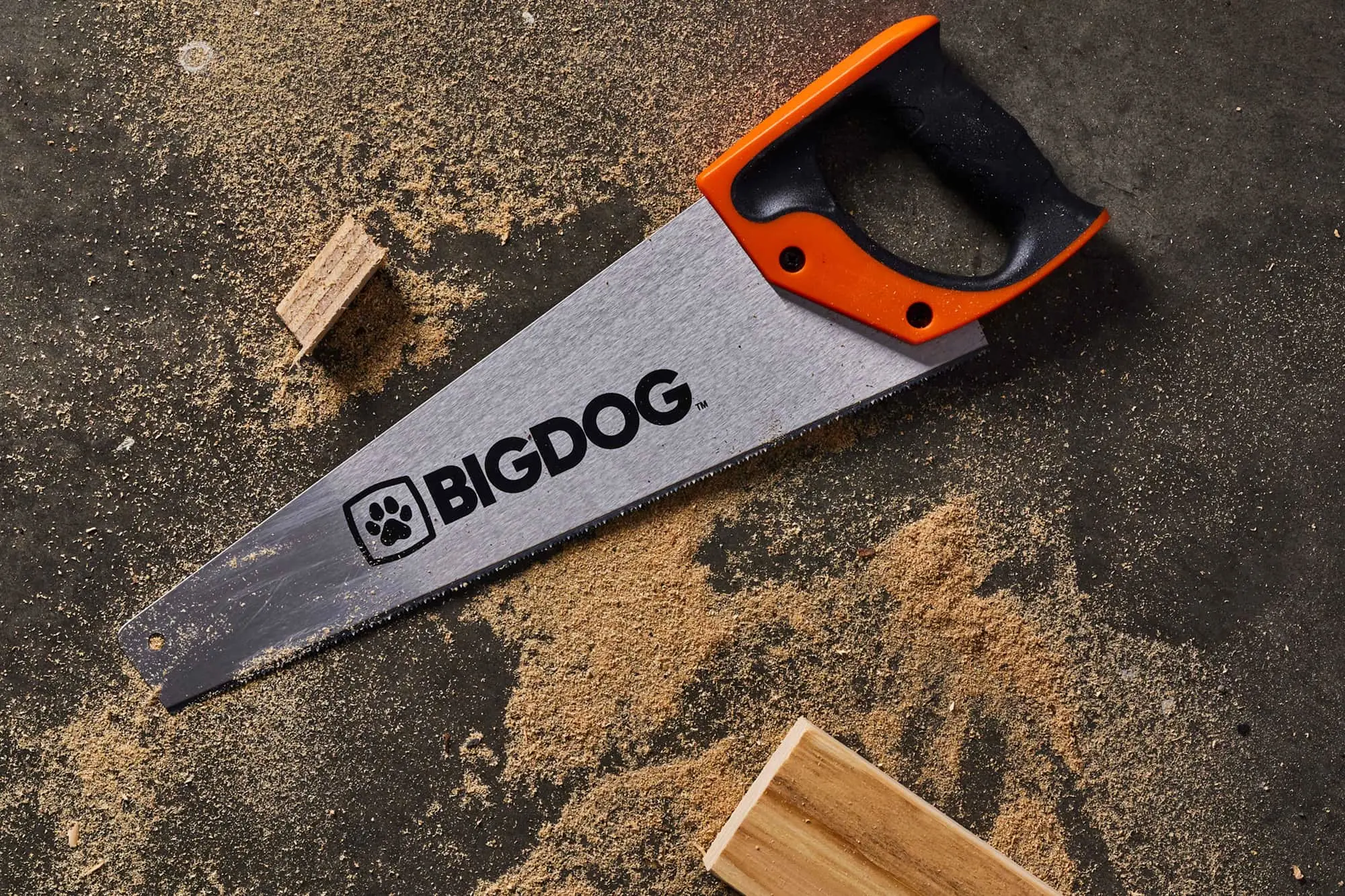 BIGDOG handsaw with orange and black handle on dark surface with sawdust and wood pieces.
