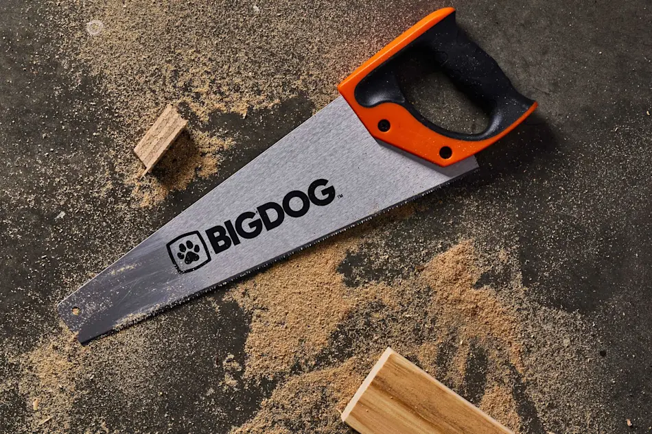 BIGDOG handsaw with orange and black handle on dark surface with sawdust and wood pieces.