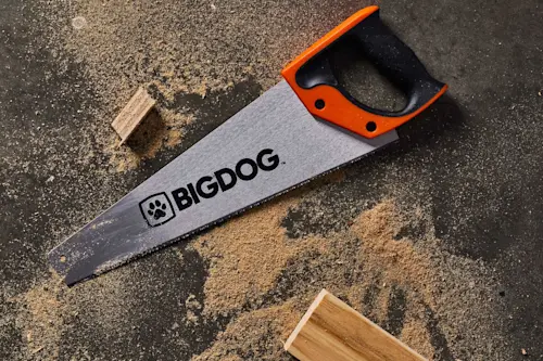 BIGDOG handsaw with orange and black handle on dark surface with sawdust and wood pieces.