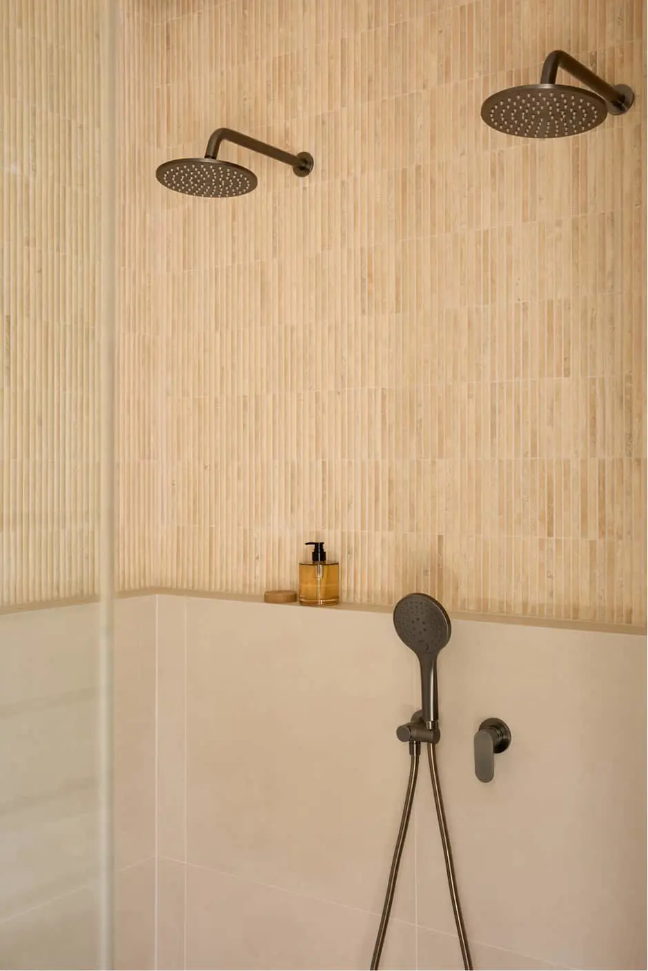 Two gunmetal rainfall showerheads mounted on a beige textured tile wall with handheld shower.