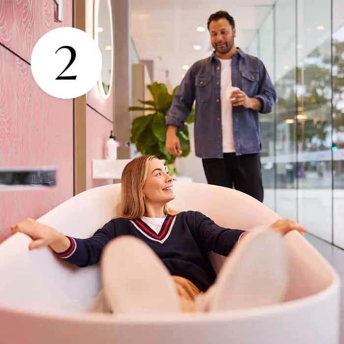 A woman sits in a pink freestanding bathtub in a showroom while a man holding a cup stands nearby.
