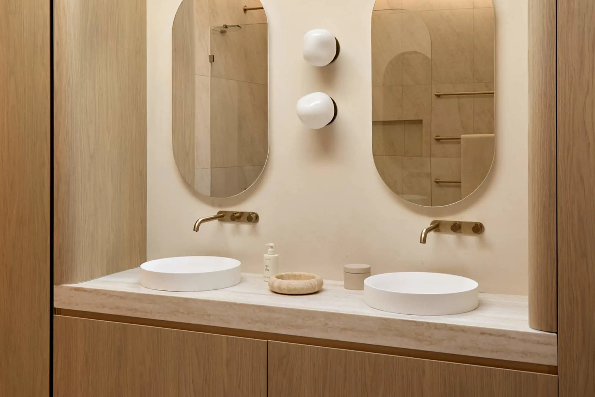 Double bathroom vanity with two white basin sinks, brass wall-mounted taps, oval mirrors, and light wood cabinetry.