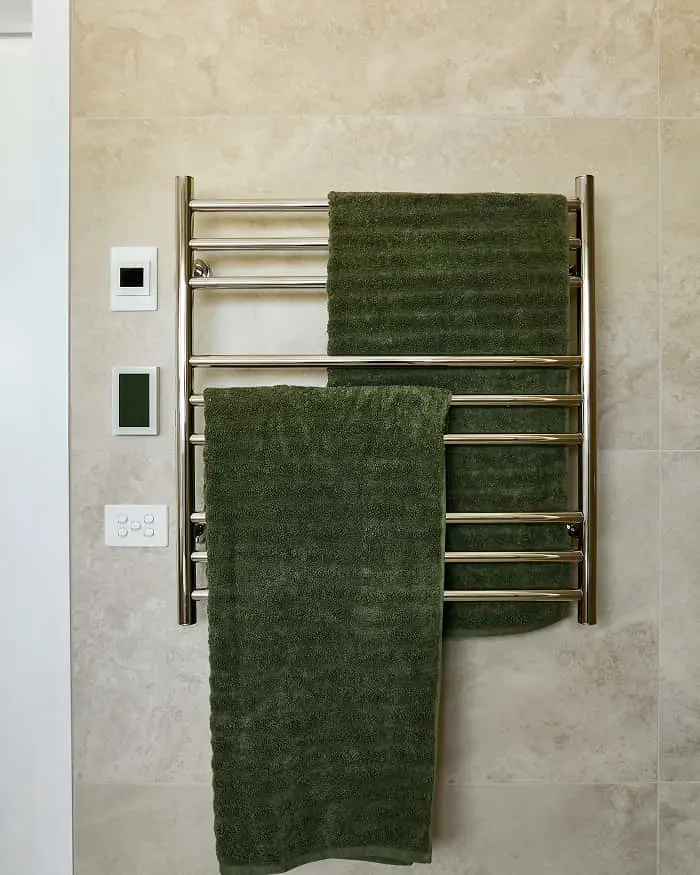 Stainless steel heated towel rack with dark green towels mounted on a beige tiled bathroom wall.