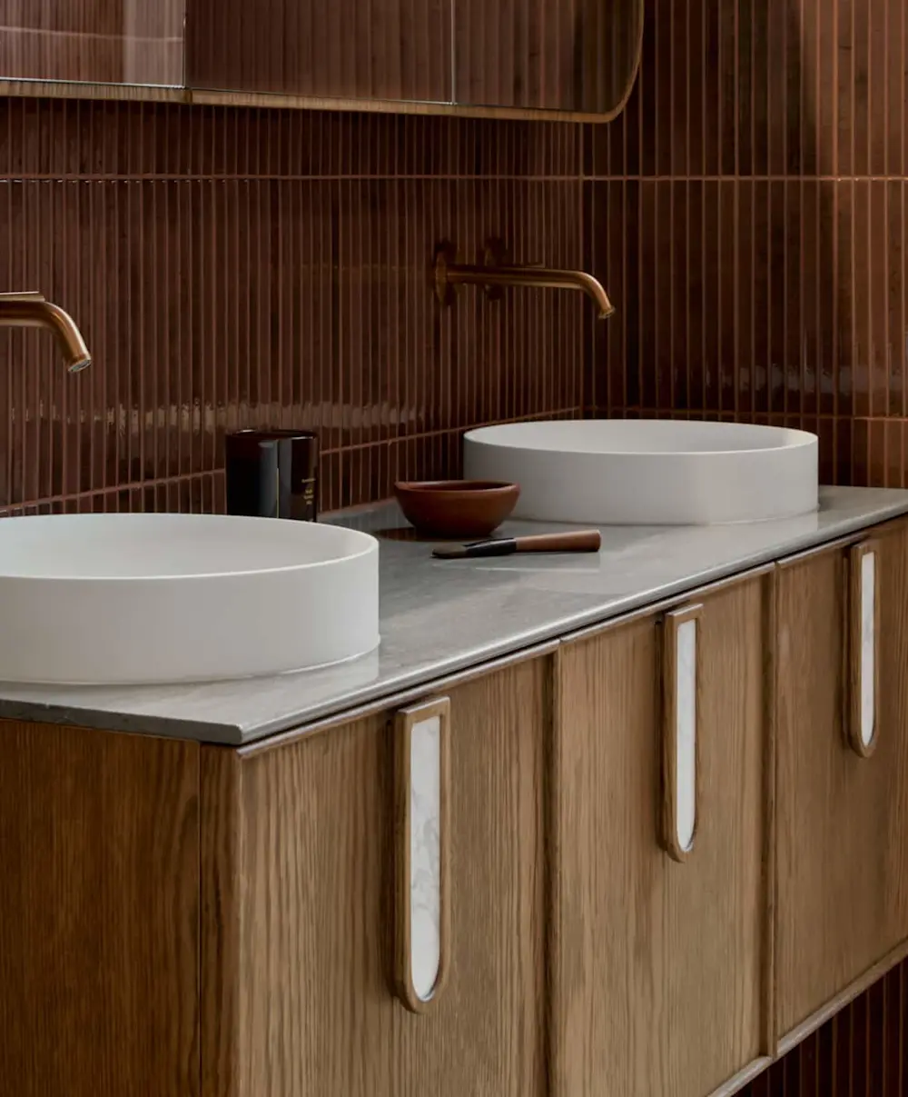 Modern bathroom with white vessel sinks on gray countertop, wooden cabinetry, and berry tiled walls with bronze fixtures.