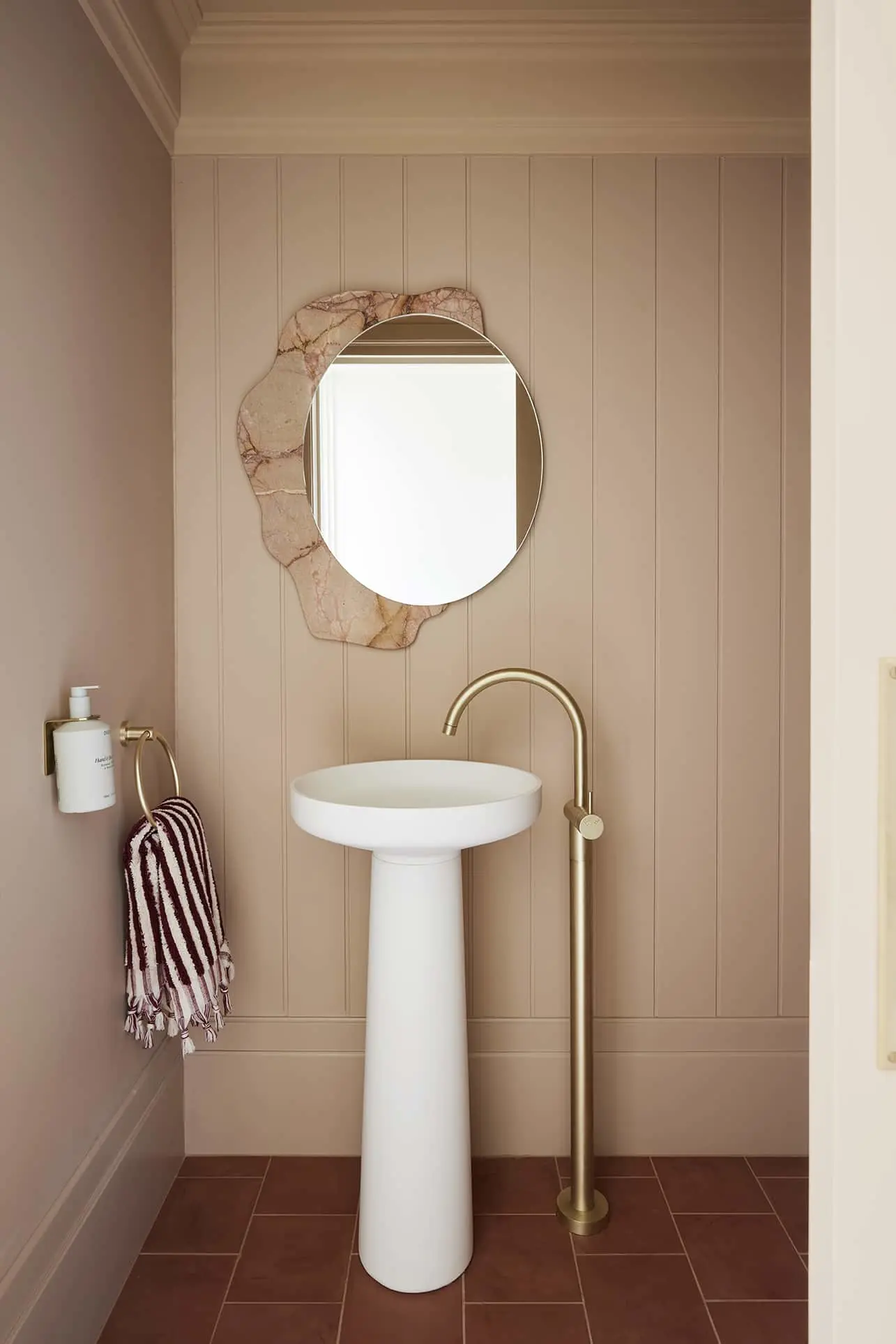 Modern bathroom with white pedestal sink, brass faucet, round mirror with marble backing, and beige paneled walls.